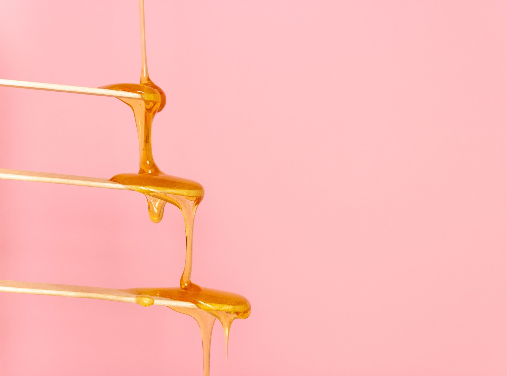 Sugar paste for hair removal runs down a wooden spatula. The concept of depilation, waxing and sugaring. Pink background, free space for text.