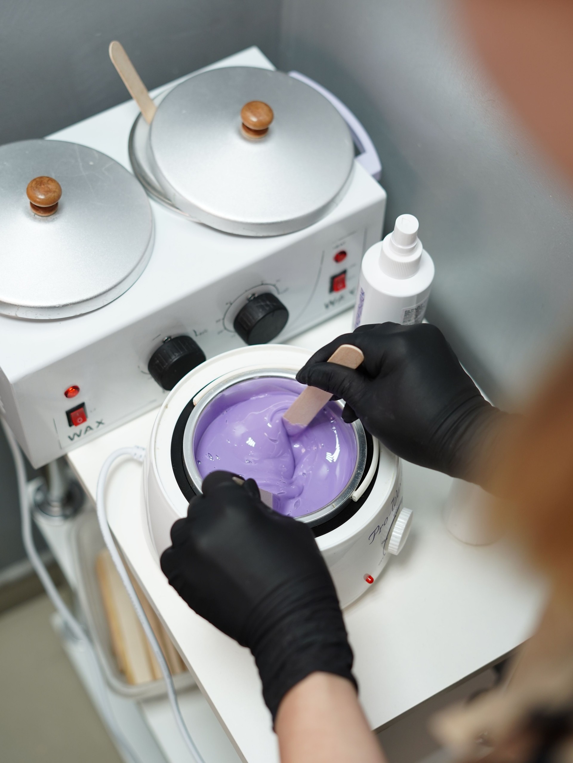 Hot violet wax used for hair removal. Close-up of depilation master hands in black protective gloves holds spatula with melted wax. Device for melting wax